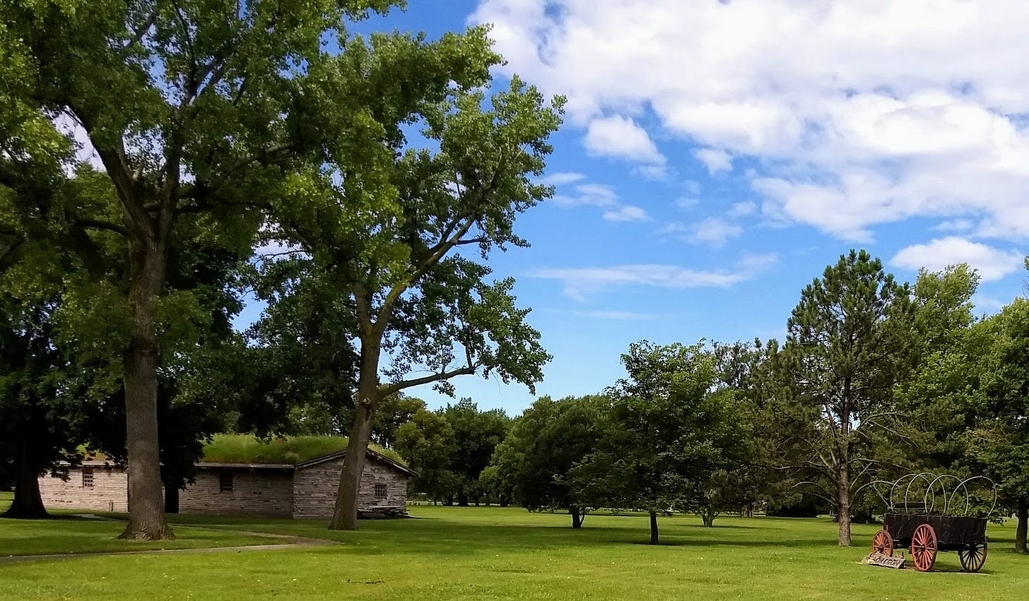 Photo of small low building with grass growing from roof and covered wagon, with grass and trees Photo of small low building with grass growing from roof and covered wagon, with grass and trees
