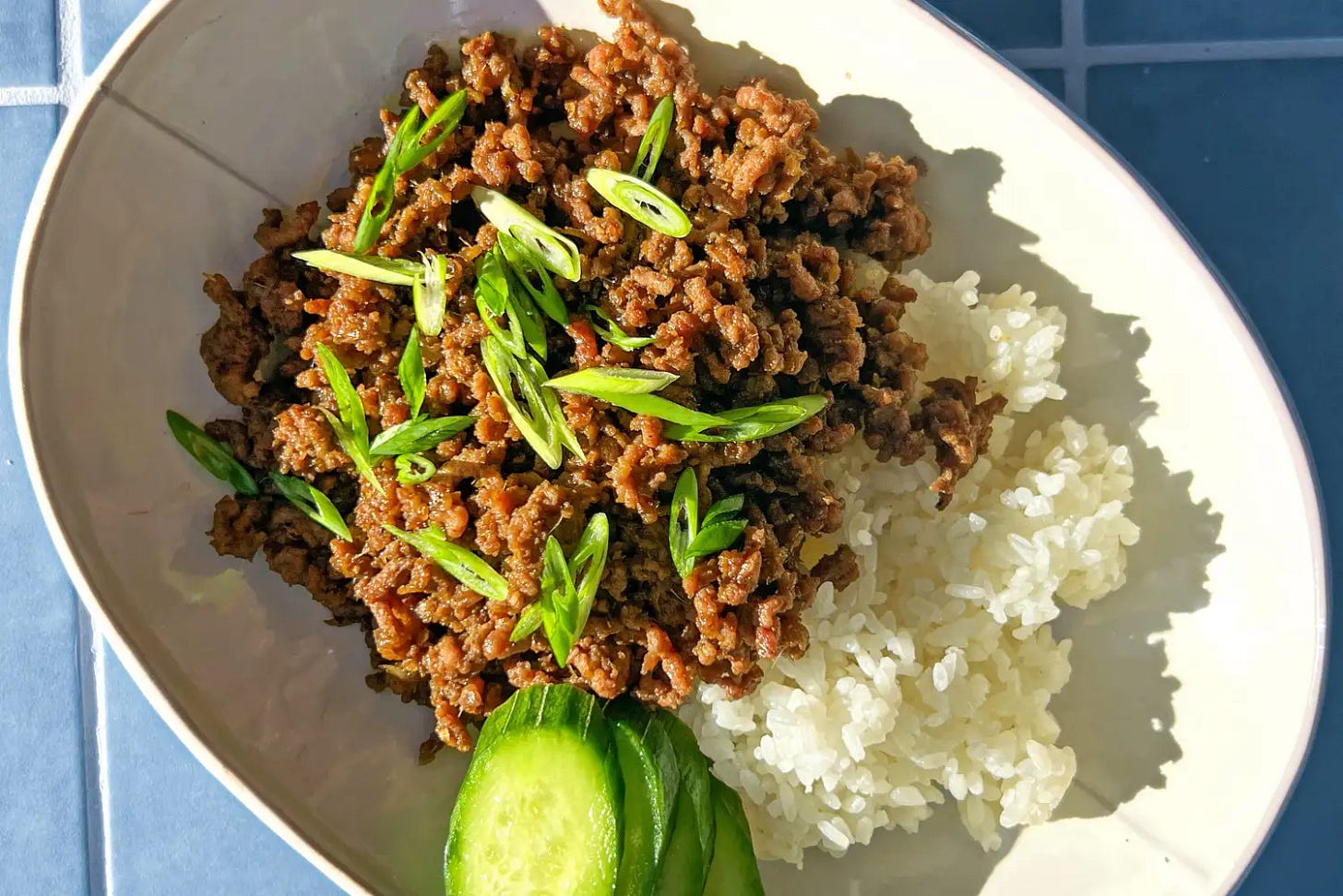 A dish with cooked ground beef served over white rice garnished with sliced scallions accompanied by cucumber slices