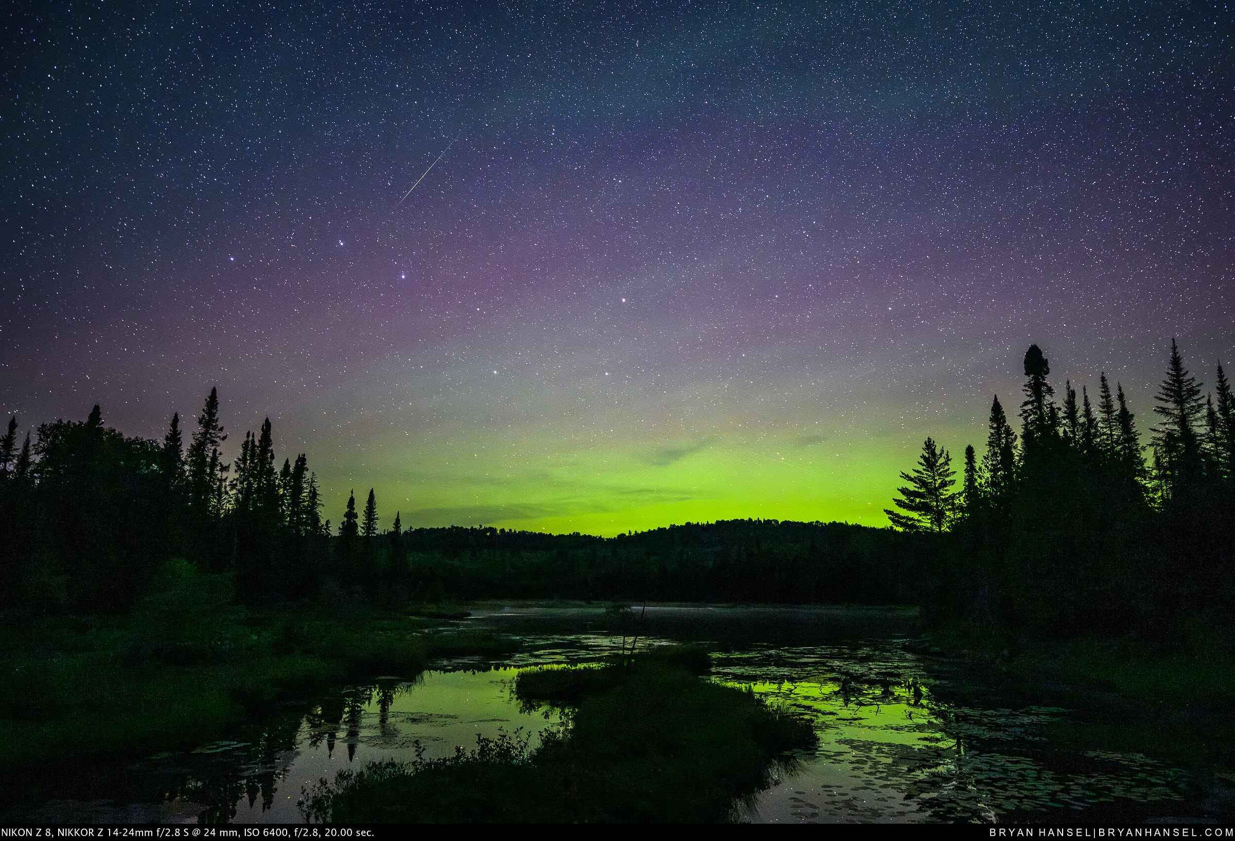 A meteor above the northern lights which reflect back into a pond with a floating bog on it.