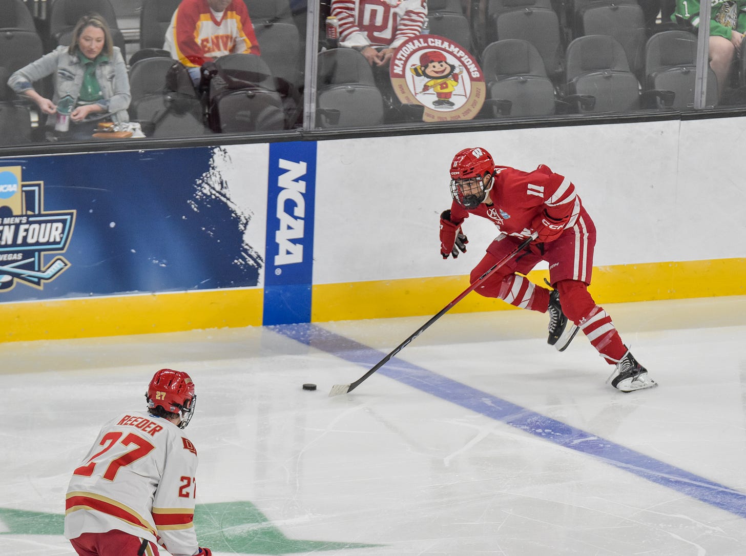 Wisconsin hockey senior Simon Tassy skates across blue line while pushing puck up ice alongside NCAA word logo.