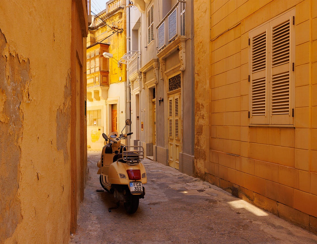 Narrow sunlit alley in Birgu, Malta, with honey-colored limestone buildings, wooden balconies, shuttered windows, and a parked cream-colored Vespa.