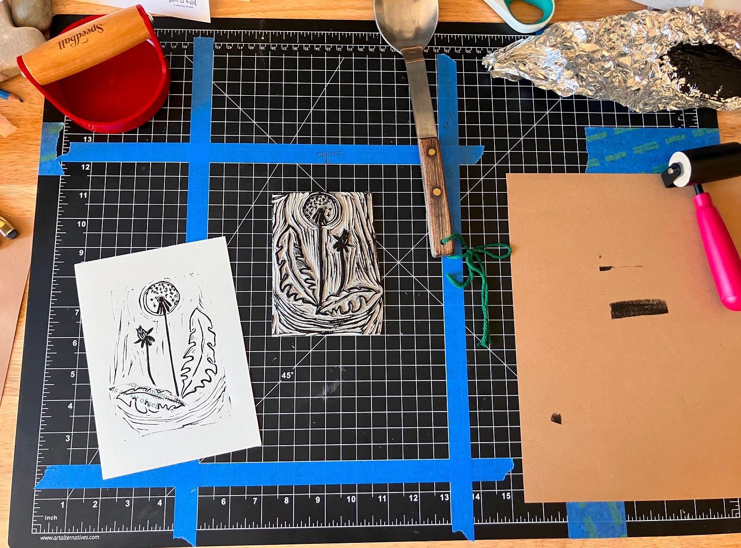 A photo of my workbench, with a black cutting mat and printmaking supplies on top. A print of a dandelion is in progress. A photo of my workbench, with a black cutting mat and printmaking supplies on top. A print of a dandelion is in progress.