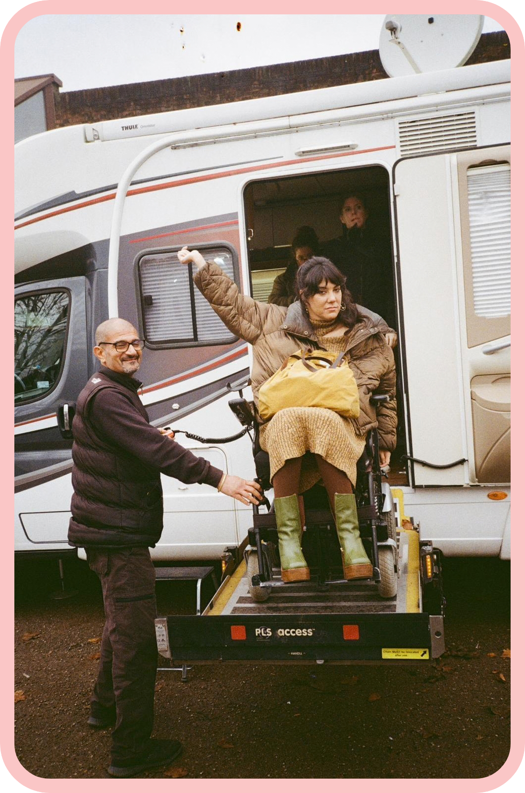 A slightly grainy analogue photo of Kyla in her wheelchair being lowered from an accessible vehicle via a platform lift. She is wearing a tan winter coat, a gold knitted dress and a yellow bag, and her expression is poker-faced but sassy, as she holds one hand raised in the air as if to say “Ta da!”. A bald man wearing black thick-framed glasses and black clothes is standing beside her and the platform lift, and his body language and gentle smile suggest he is providing assistance.