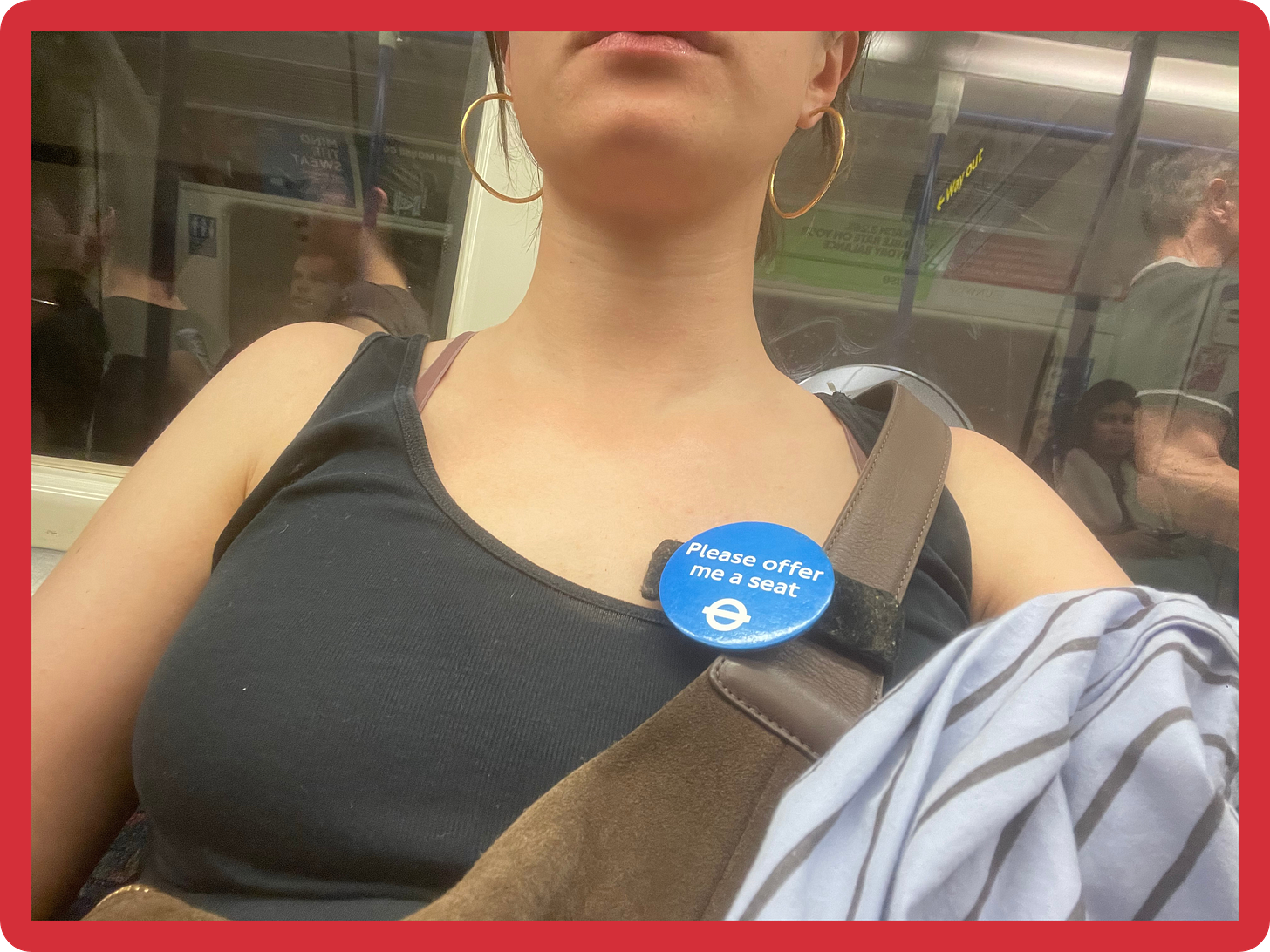 A selfie, taken from a low-angle, of a young white woman whose face is not visible, sitting on a crowded Tube and wearing a blue 'Please offer me a seat' badge, which has been attached to her brown suede crossbody bag. She is wearing gold hoop earrings, and holds a blue and black striped shirt over her shoulder. A selfie, taken from a low-angle, of a young white woman whose face is not visible, sitting on a crowded Tube and wearing a blue 'Please offer me a seat' badge, which has been attached to her brown suede crossbody bag. She is wearing gold hoop earrings, and holds a blue and black striped shirt over her shoulder.