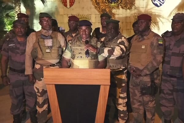 Several men in military uniform stand around a podium as one man holds a microphone for another. Several men in military uniform stand around a podium as one man holds a microphone for another.