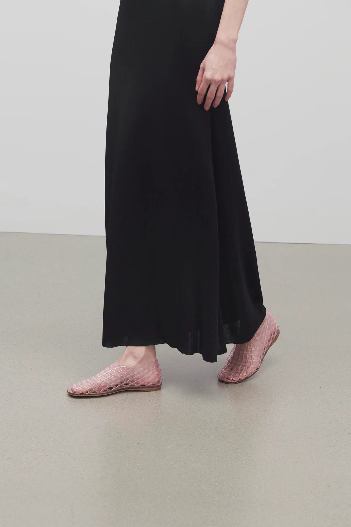 A cropped image depicts the lower body of a woman wearing an ankle-length black skirt and clear flat shoes made from PVC in lattice pattern.