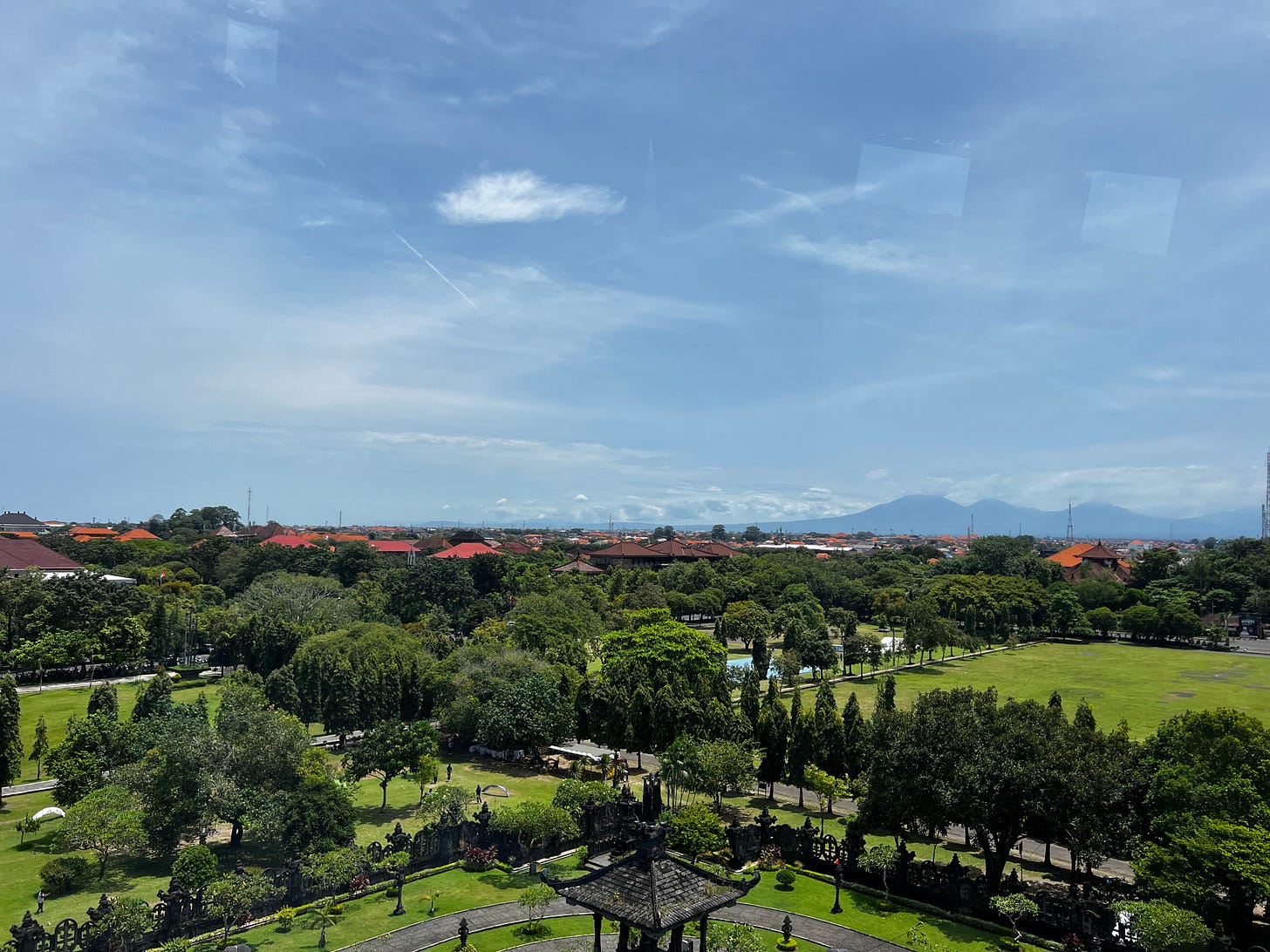 Aerial view over Bali: a green park and temple-like pavilion in the foreground, red-roofed buildings beyond, and mountains on the horizon beneath a wide blue sky. Aerial view over Bali: a green park and temple-like pavilion in the foreground, red-roofed buildings beyond, and mountains on the horizon beneath a wide blue sky.