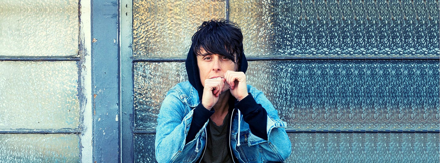 An image of Andrea in a black hoodie and jean jacket. They are holding their hands up near their face. In the background is a big, weathered window.