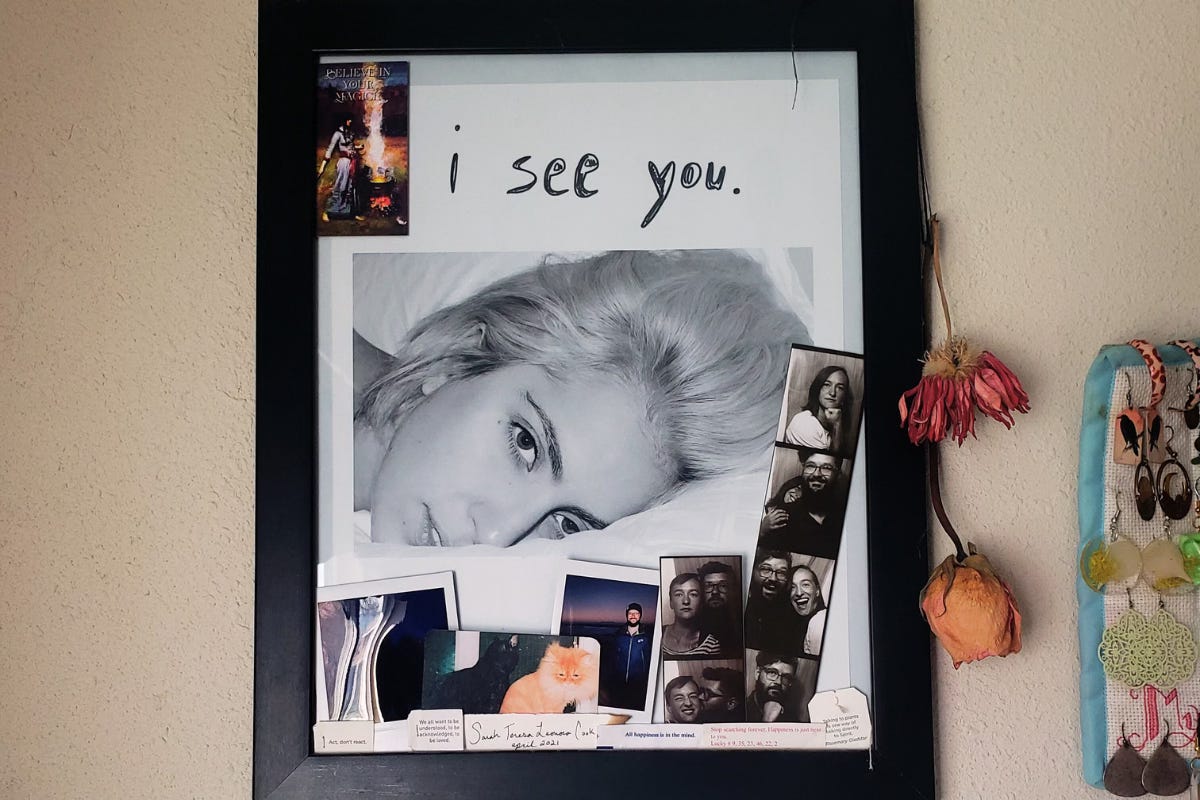 A close-up black and white photograph of Lady Gaga's face is hung on a wall in a black frame. "I see you" is written in Sharpie right above her face. Other ephemera is tucked into and hanging of off the frame, including dried flowers, instant photos, and some photobooth strips that show images of Sarah and M.