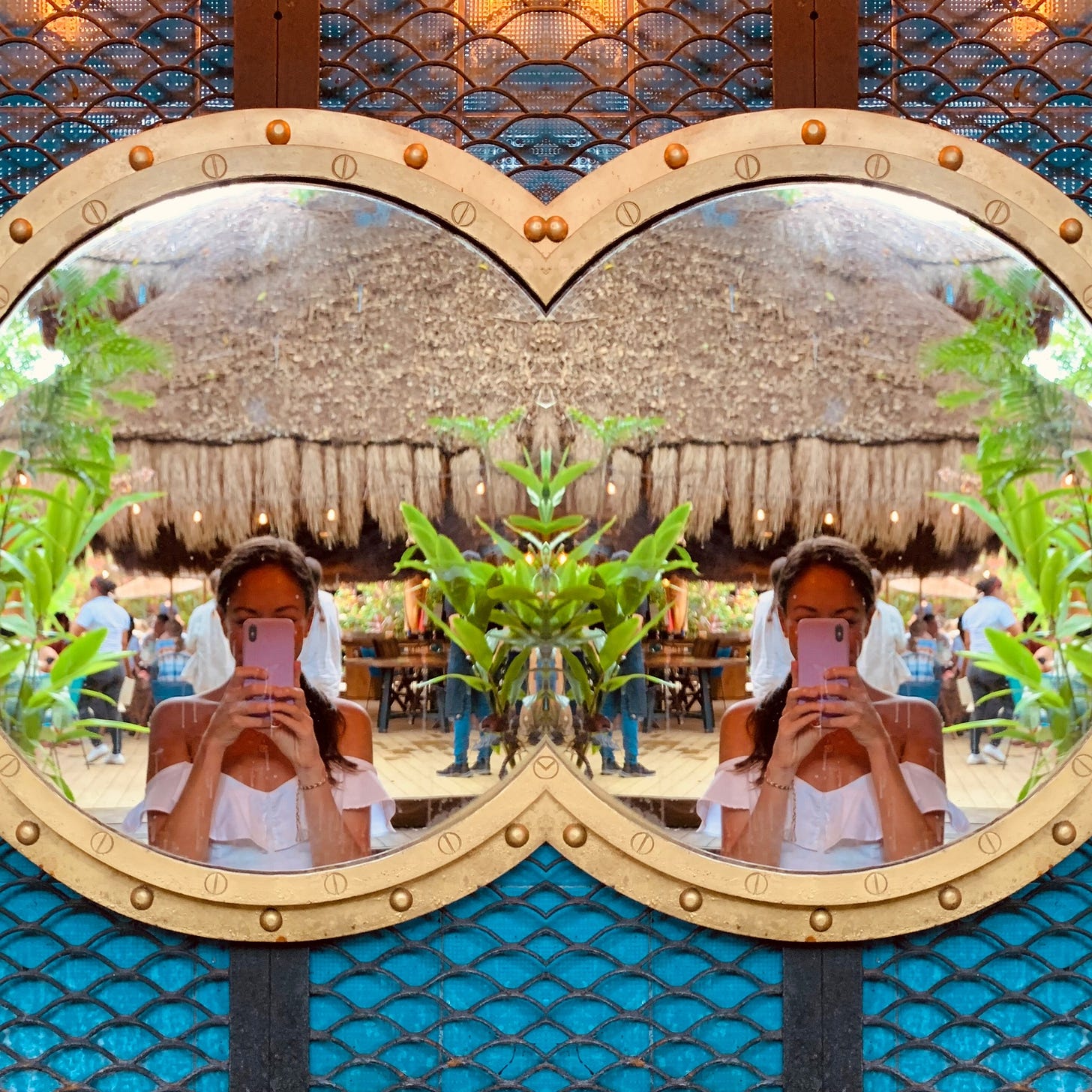 A woman in a white top takes a mirror selfie in a heart-shaped frame that creates a symmetrical reflection, visually duplicating her and the tropical restaurant behind her. The image evokes themes of identity, self-reflection, and duality.