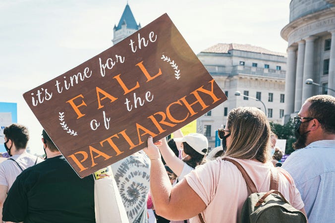 Male and female demonstrators. One of the women has a sign that reads "It's time for the fall of the Patriarchy."