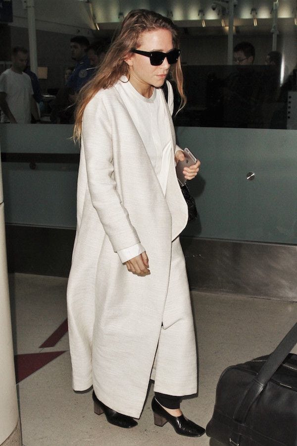 This may contain: a woman in white coat and sunglasses walking through an airport