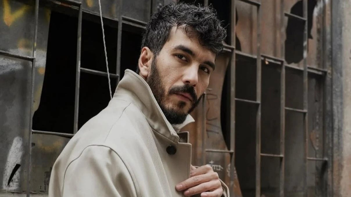 Mert Doğan oyuncu fotoğrafı — bej palto ile endüstriyel arka planda