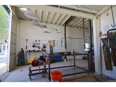 One row of three photos, left to right: Photo of two chairs and a rectangular wooden table topped with paint cans, papers, and a book, backed by a hanging frame of colorful artwork and a black and dark green stone fireplace; Photo of the high-ceilinged garage studio workshop filled with buckets, cans, tools, including for welding, and a large metal sculpture hanging from the rafters; Photo of a woman and man, both laughing and wearing hats, lay on lounge chairs in the sun with books at their sides, next to a large white wooden bird house with tucked-in bird feeder.
