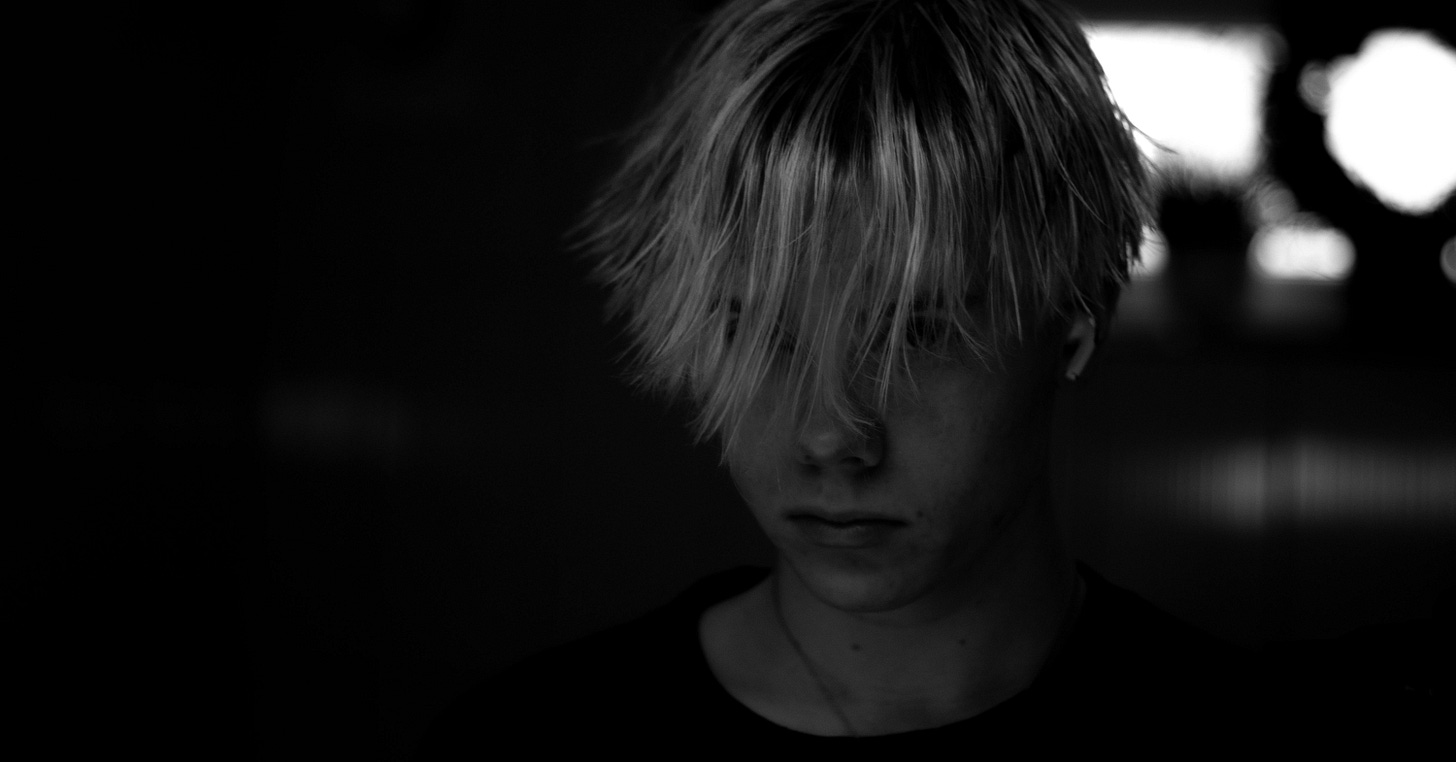 A close-up of a person with messy, short blonde hair standing in a dimly lit room with a blurred background and soft lighting.