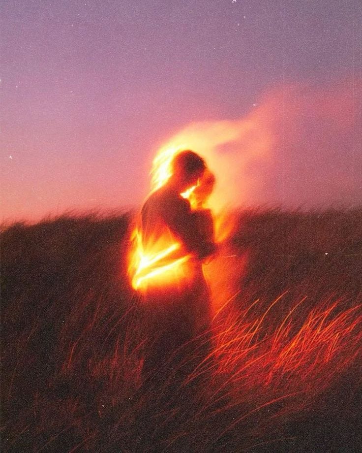 This may contain: a person standing in the grass with their arms around each other and glowing fire behind them