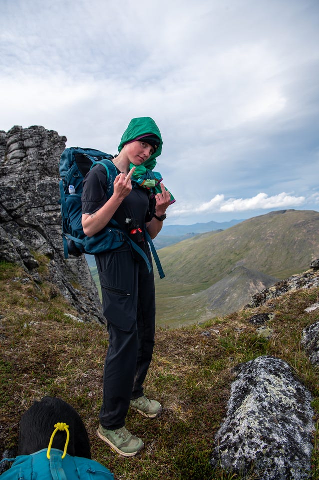 A hiker with a teal pack stands on a tundra ridge raising her middle fingers toward the camera, a green wind jacket wrapped and tied around her head as a hood. A dog pack sits in the foreground, and pale grassy ridges roll away under a bright, cloudy sky.