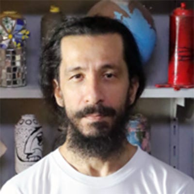 Homem com cabelos escuros, de comprimento médio, penteados para trás, barba e bigode cheios. Veste uma camiseta branca e calça jeans clara. O braço esquerdo é coberto por tatuagens geométricas coloridas. Ao fundo, há uma grande estante com latas de tinta spray utilizadas como peças artísticas e decorativas. Homem com cabelos escuros, de comprimento médio, penteados para trás, barba e bigode cheios. Veste uma camiseta branca e calça jeans clara. O braço esquerdo é coberto por tatuagens geométricas coloridas. Ao fundo, há uma grande estante com latas de tinta spray utilizadas como peças artísticas e decorativas.