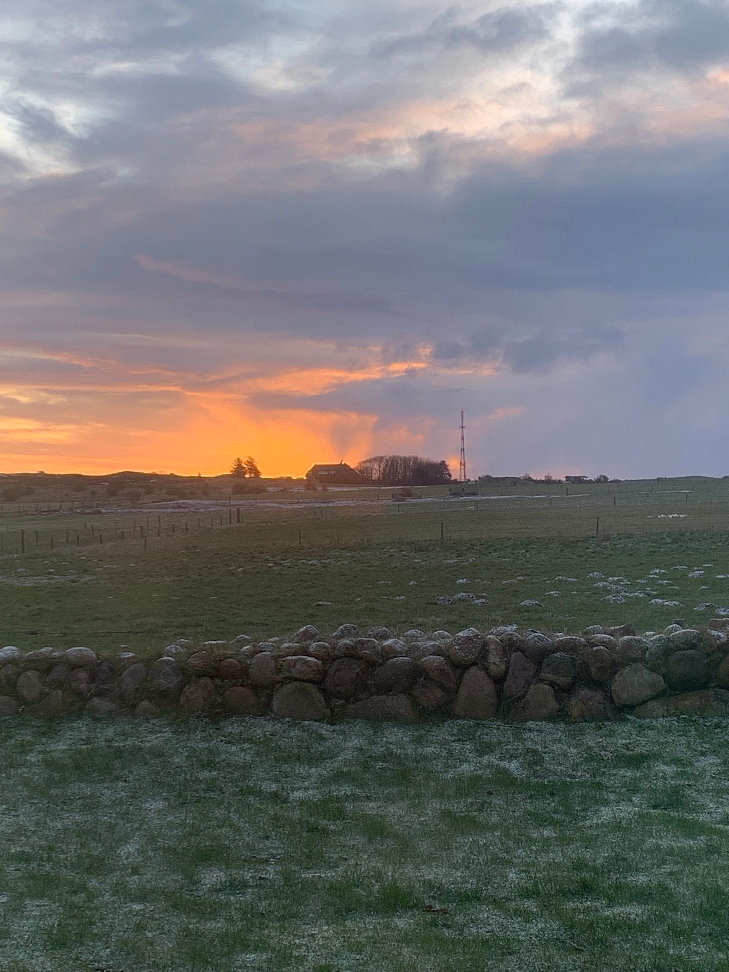 A grey sky on the top of the frame. It is sunset, there is a red splash of light falling onto a house off in the distance. A field in dusky colors makes up the bottom half of the frame. 