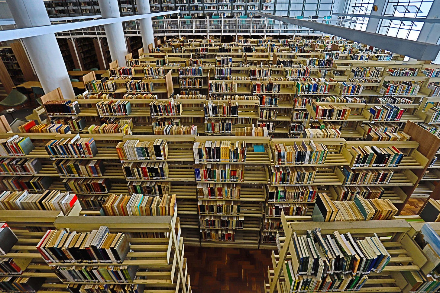 #descrevemospravocê  A imagem mostra o interior amplo de uma biblioteca moderna, vista de cima. O espaço é preenchido por dezenas de estantes longas e alinhadas, repletas de livros organizados em fileiras. As estantes são feitas de madeira clara e têm estrutura vazada, o que permite visualizar parcialmente os livros de diferentes ângulos.  Ao fundo, há colunas brancas e janelas de vidro que deixam entrar luz natural, iluminando o ambiente de forma suave. No andar superior, pode-se ver uma passarela com guarda-corpo branco, que percorre parte do mezanino onde também há estantes e mais livros. Cadeiras verdes e mesas de estudo estão posicionadas entre as colunas, sugerindo um espaço de leitura silencioso e convidativo.  O piso é de madeira escura, contrastando com o tom claro das estantes. A imagem transmite uma atmosfera de organização, conhecimento e tranquilidade, típica de bibliotecas públicas ou universitárias.