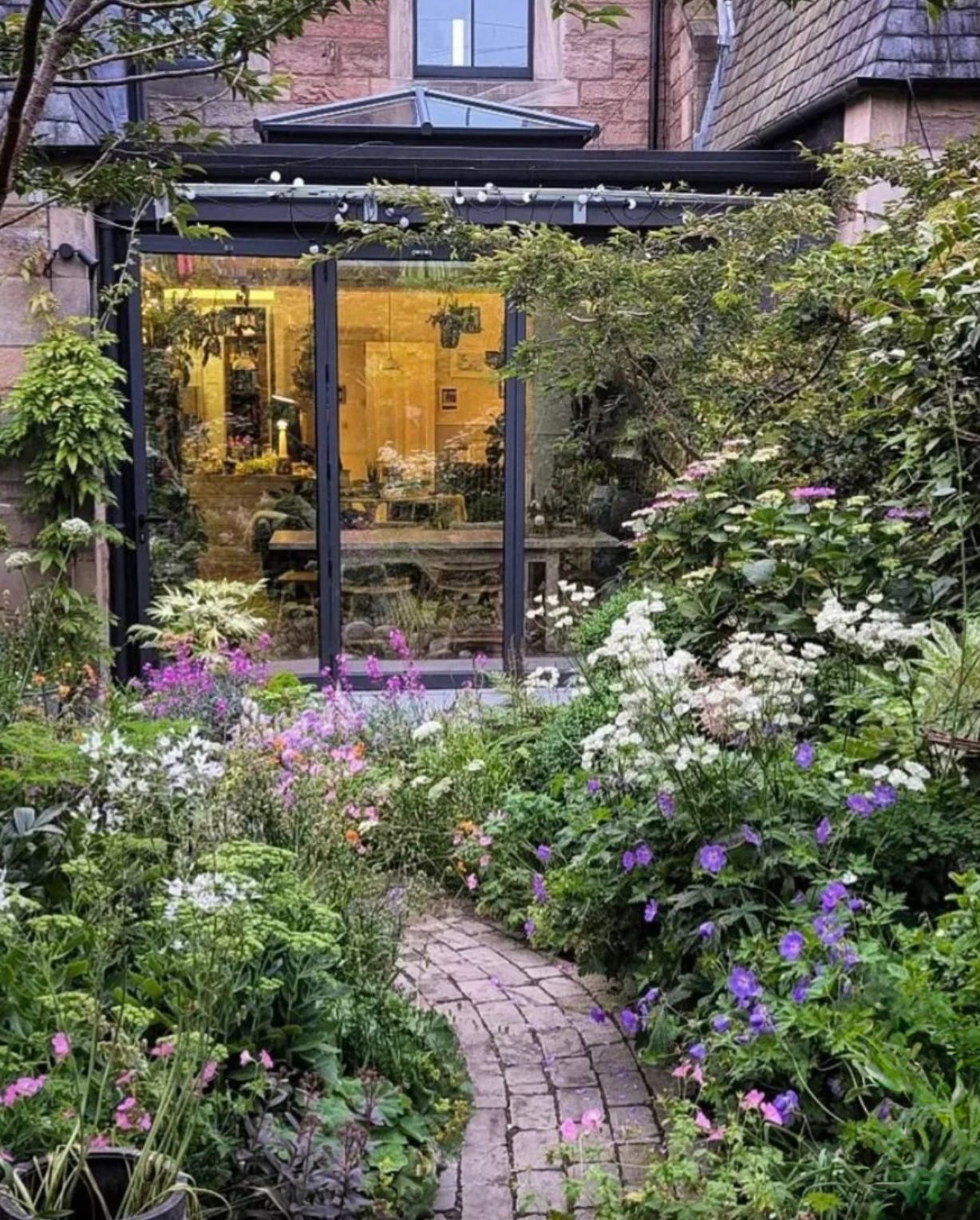 Colourful cottage garden with mixed flowering borders and a winding brick pathway leading to a contemporary glass patio room.