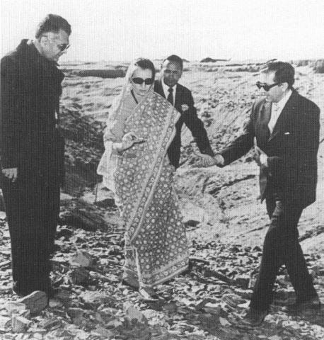 Indira Gandhi, in a patterned sari, walks across broken desert ground with three men in dark suits at the Pokhran test site