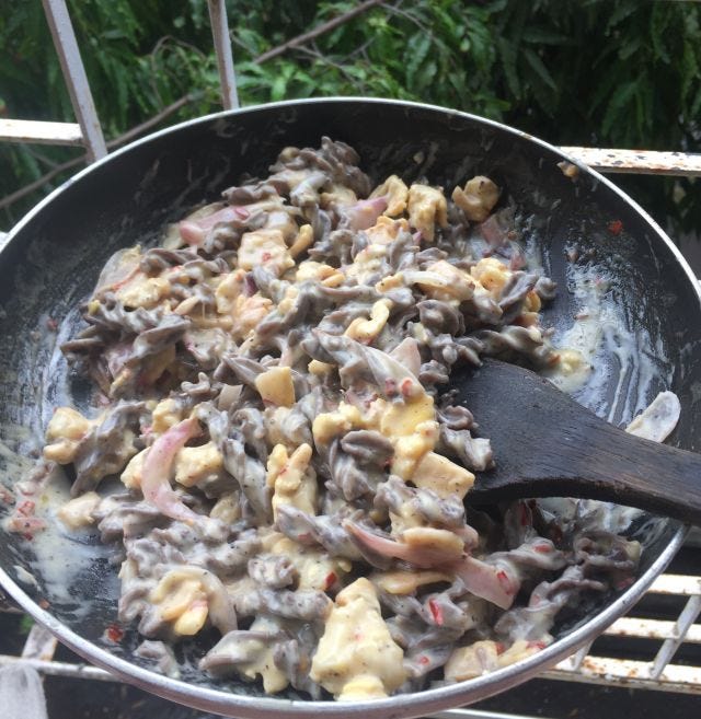 A pan with black fussili pasta, eggs, capsicum, mushroom in white sauce with a ladle.
