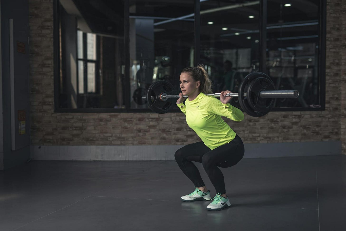 Woman in a neon green top squats with a barbell in a gym. Focused expression. Brick wall and mirrored windows in the background.