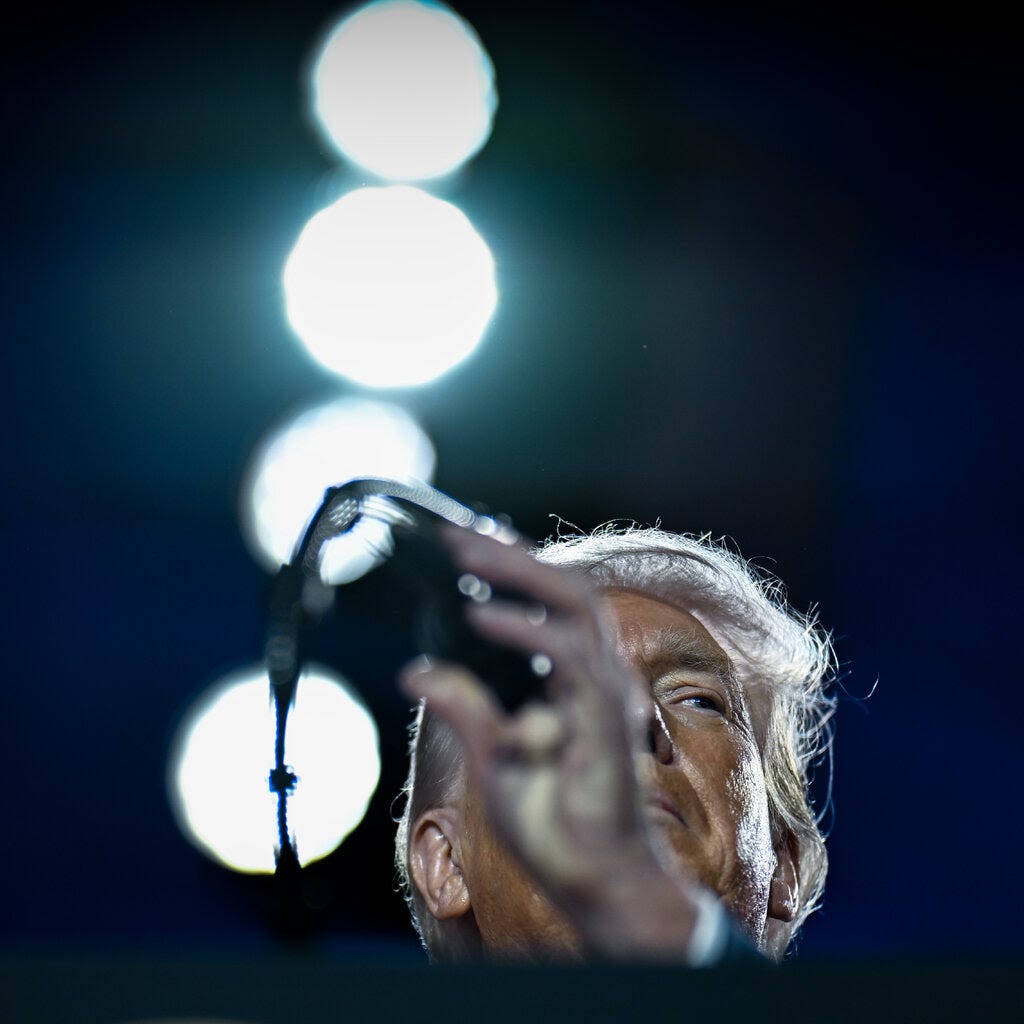President Trump gesturing, with a microphone in front of him and four lights puncturing a dark background behind him.