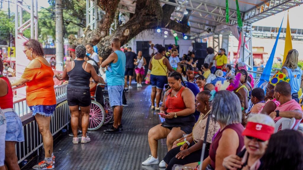 #PraTodosVerem: A imagem mostra um camarote acessível durante o Carnaval, com diversas pessoas idosas e com deficiência. O espaço é coberto e conta com uma estrutura metálica. Algumas pessoas estão sentadas em cadeiras brancas, enquanto outras estão em pé, em cadeiras de rodas ou apoiadas nas grades que cercam o local. 