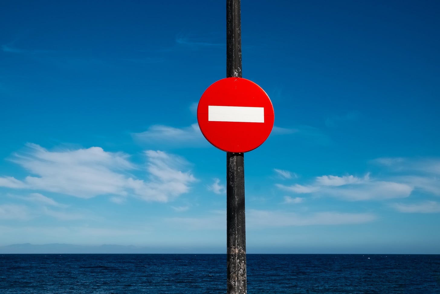 Sfondo azzurro di cielo e mare.In primo piano un palo con il cartello di divieto d'accesso con cerchio rosso e linea bianca orizzontale.