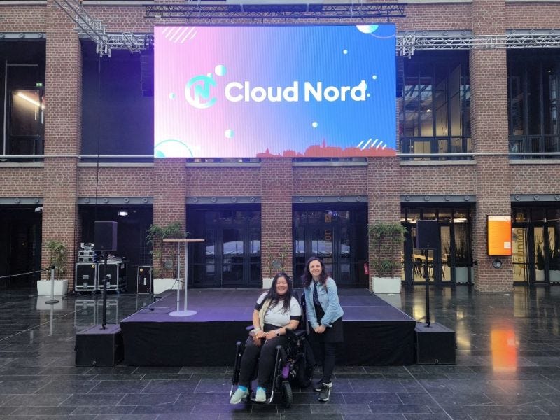 Dans une grande salle avec des murs en briques, devant une scène, Céline, jeune femme brune portant une veste en jean, sourit à côté de Thanh, jeune femme brune portant un haut blanc. Derrière elle, un écran affiche “Cloud Nord”.