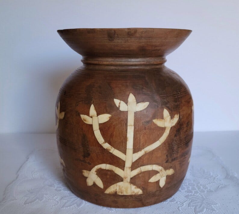 May include: A brown wooden vase with a white floral design. The vase has a wide mouth and a rounded body. The floral design is made up of small white pieces that are inlaid into the wood.