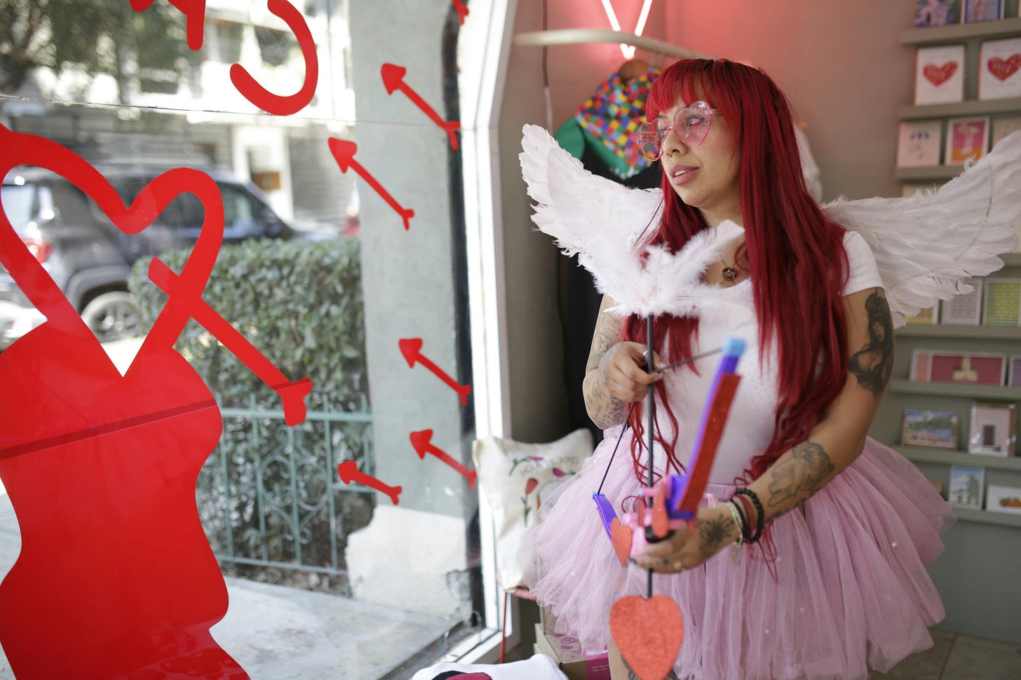 Woman with red hair and tattoos in a cupid costume looking out a window from a Valentine's Day display.