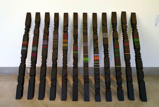 Monumental installation of tall vertical redwood elements with charred surfaces and bands of bright sari thread, arranged in a row against a white gallery wall. Monumental installation of tall vertical redwood elements with charred surfaces and bands of bright sari thread, arranged in a row against a white gallery wall.