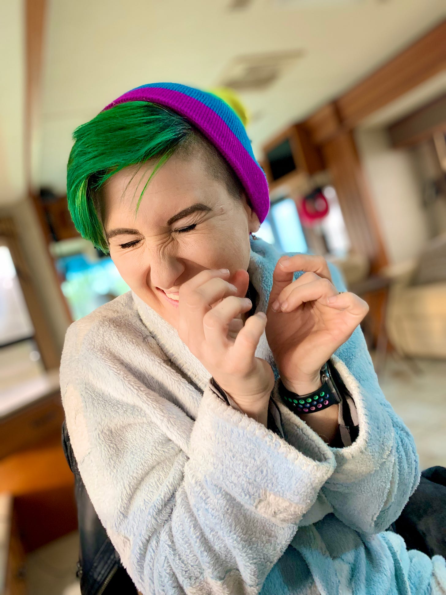 Photo of Lyric, a fair skinned nonbinary human with short green hair in a blue cloud bathrobe and a rainbow beanine, happy stimming with their hands near their face