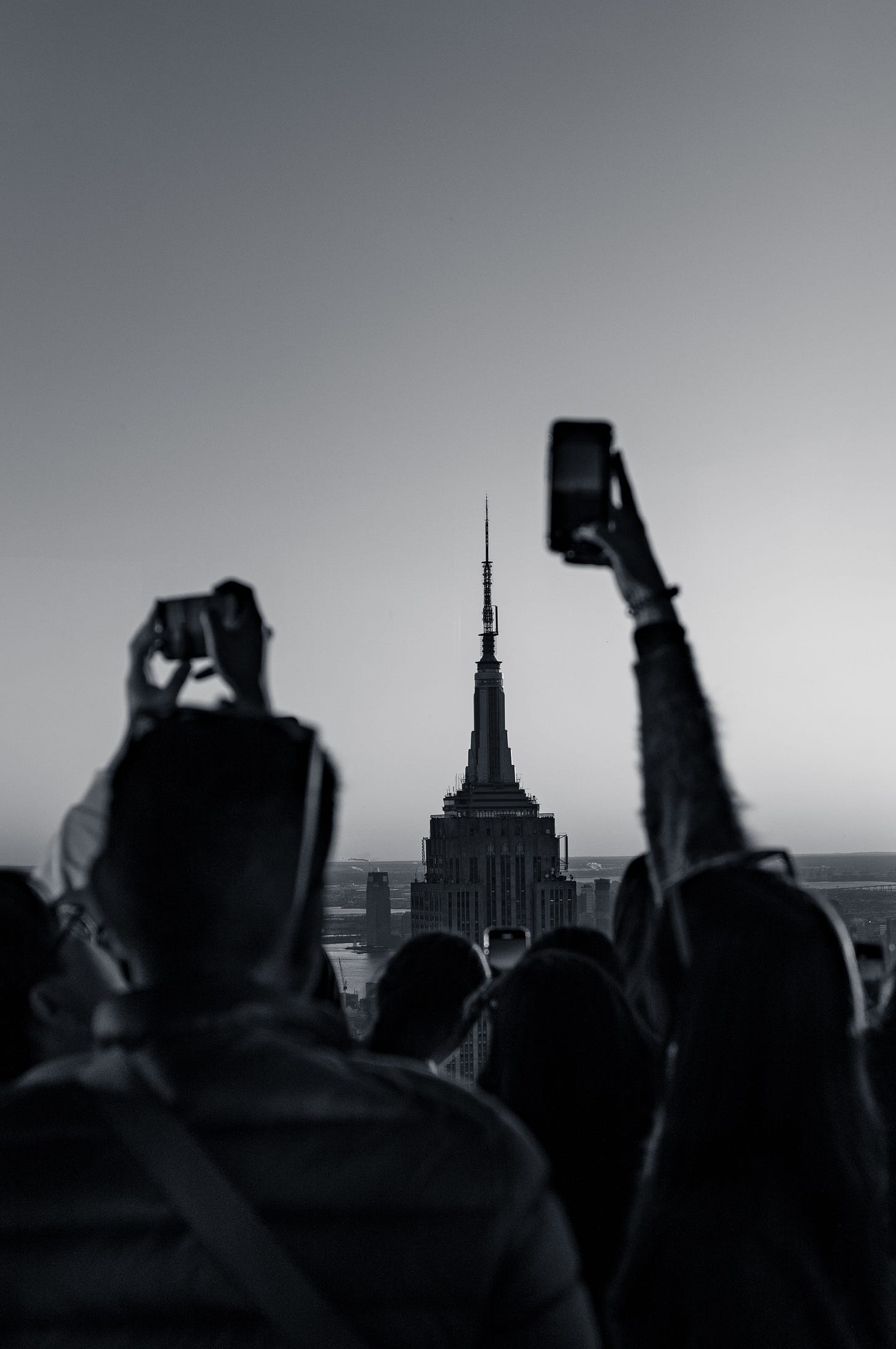 A crowd seen from behind with phones raised at Summit One Vanderbilt, the spire of the Empire State Building centred between them, New York, in black and white.