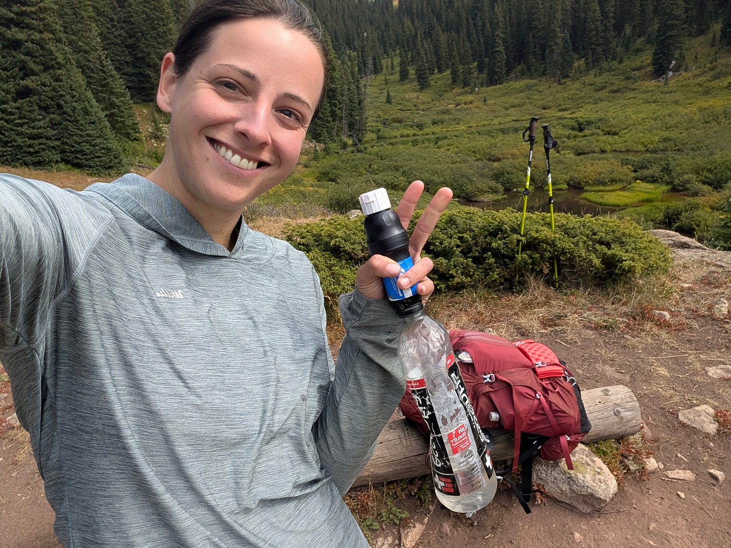 Smiling hiker taking a trail break with a backpack and water filter, seated near a stream in a forested mountain landscape.
