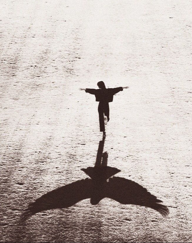 A black-and-white photograph of a child running with arms outstretched on a wide, empty surface. Their shadow on the ground forms the shape of a large bird with spread wings, creating a surreal visual effect.