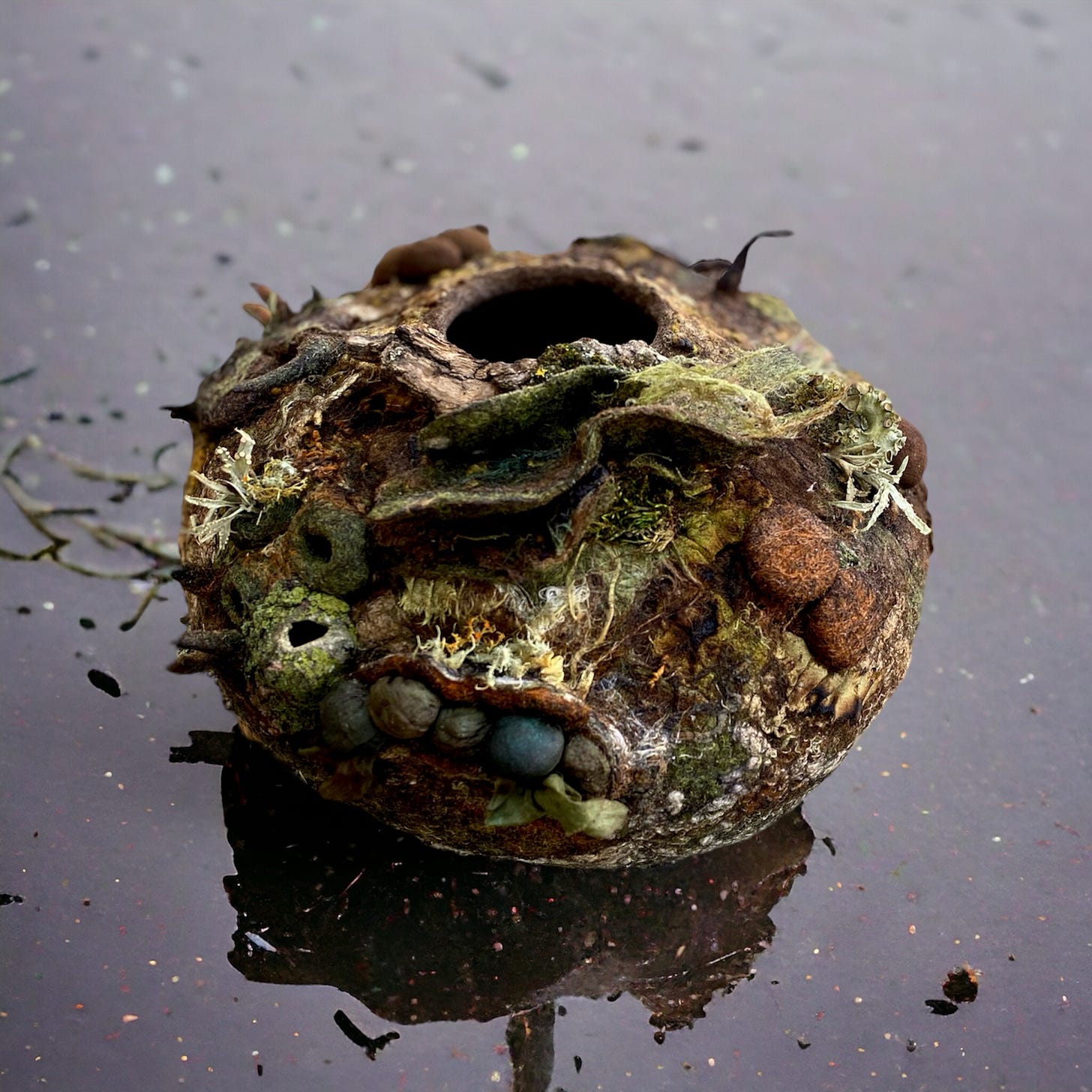 A textured, organic-looking felt vessel with mossy and stone-like details, reflecting on a wet surface.