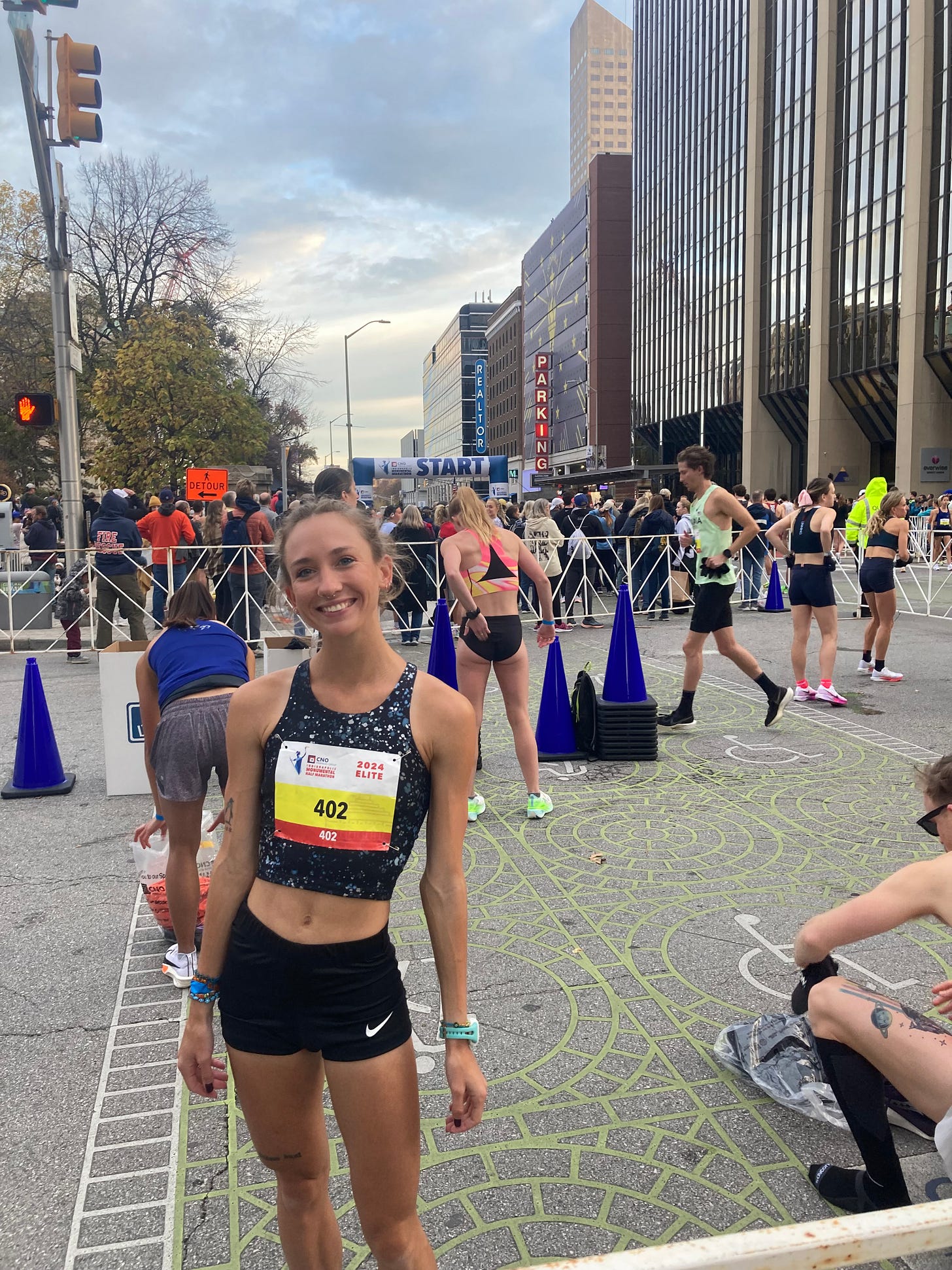 Woman stands with race bib near the start line of the Indianapolis Monumental Half Marathon. She wears Nike shorts and a Garmin Watch. Her competitors stand behind her. She is in the elite athlete field.