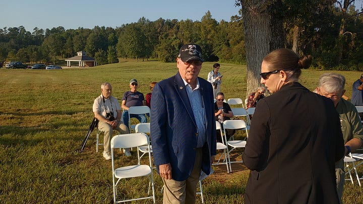A group of veterans and community members pose outdoors at a POW*MIA memorial. In the foreground is a black granite monument engraved with the POW*MIA emblem and the words “YOU ARE NOT FORGOTTEN,” with a small American flag at its base. Several U.S. flags stand behind the group. Some people wear military dress uniforms and VFW-style caps; one cap reads “Tallahassee,” and another hat bears the U.S. Air Force insignia. On the right, an elderly veteran in a wheelchair salutes while a man in a suit speaks with him. The scene is on a sunny day in a grassy field with cars and flagpoles in the background.