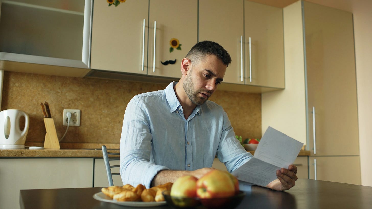 A man sits at a desk holding a piece of paper with a concerned expression, suggesting financial stress or review of bills. Photo by Vitaly Gariev on Unsplash