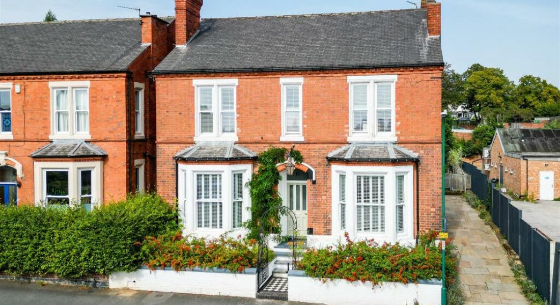 A large brick house with white windows, a white border with hedges, and a black gate, with a house next to it A large brick house with white windows, a white border with hedges, and a black gate, with a house next to it