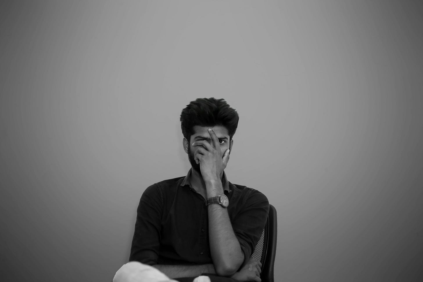Black and white portrait of a frustrated entrepreneur covering part of his face with hand, wearing dark shirt and wristwatch, sitting against plain wall - perfect visual representing business planning overwhelm and entrepreneurial analysis paralysis