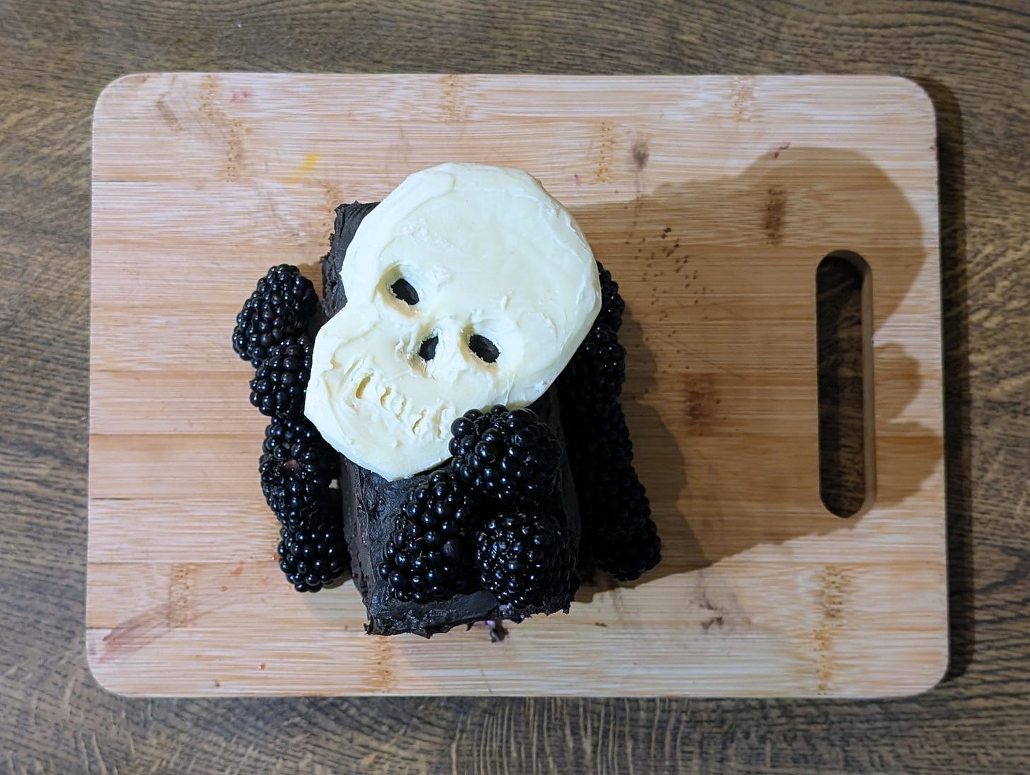 A rectangular chocolate cake rests on a wooden board, its surface coated in dark, glossy icing. Plump blackberries cluster along the edges and corners, their deep purple nearly blending into the chocolate. A pale white skull made of icing or fondant sits centered on top, creating a stark, gothic contrast against the rich, moody colours.