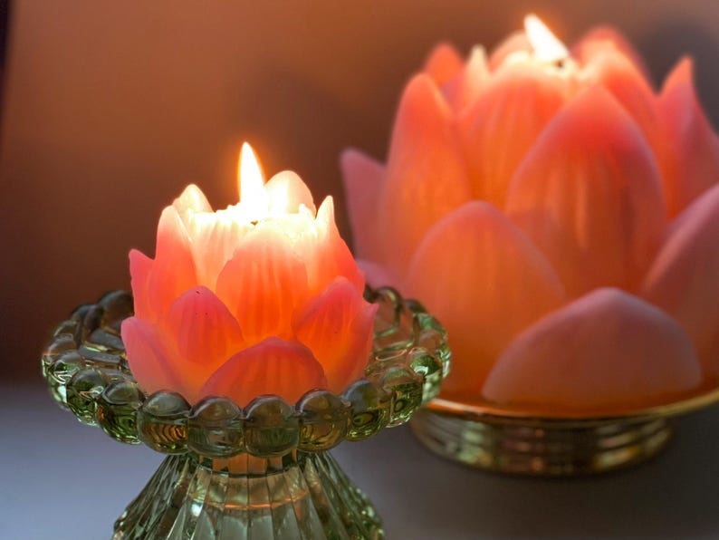 May include: Two lotus-shaped candles, one smaller and one larger, are shown lit.  The smaller candle sits on a green glass pedestal. The larger candle rests in a gold-colored dish. Both candles are a soft peachy-pink color and have a realistic lotus flower design.  The candles are illuminated, creating a warm, ambient glow.