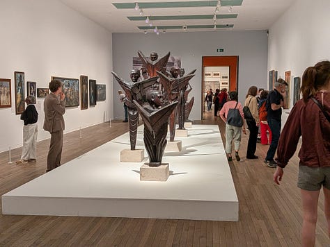 pictures taken at the gallery. Image 1 is of a door with carvings of people. Image 2: a wide picture of the galleries with wooden sculptures. Image 3: An image of a Nigerian sculptor at work in Buckingham Palace