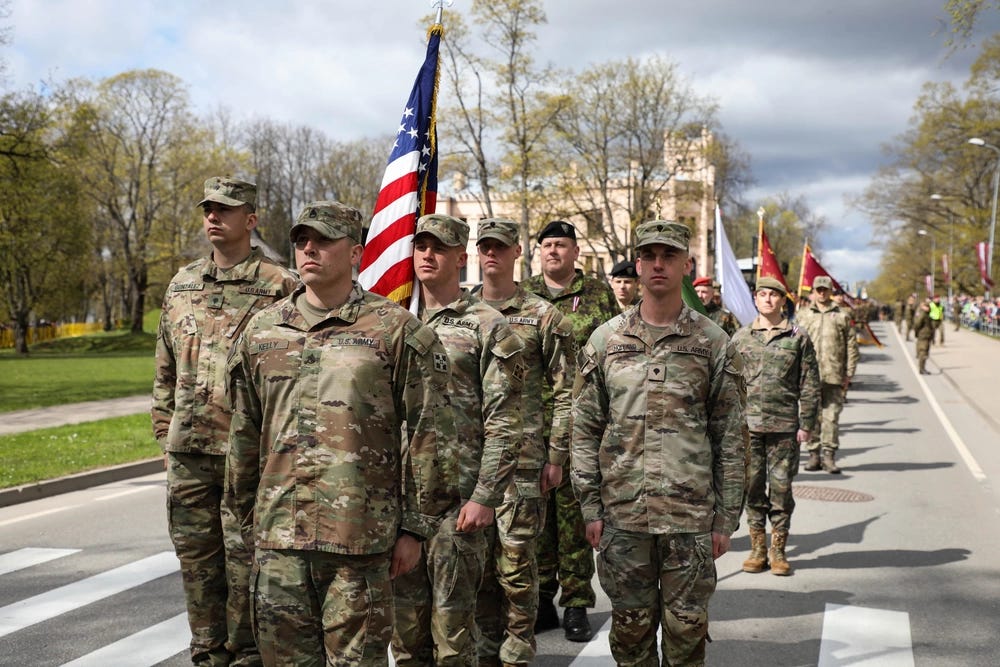 A formation of U.S. Army soldiers in MultiCam/OCP camouflage uniforms marches in a military parade, carrying the American flag. The procession takes place on a paved road lined with trees and spectators, with a classical-style building visible in the background. Additional military personnel from what appears to be at least one other nation's armed forces follow behind the U.S. contingent. A formation of U.S. Army soldiers in MultiCam/OCP camouflage uniforms marches in a military parade, carrying the American flag. The procession takes place on a paved road lined with trees and spectators, with a classical-style building visible in the background. Additional military personnel from what appears to be at least one other nation's armed forces follow behind the U.S. contingent.