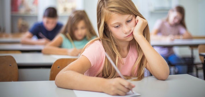 KO-mid-class-slump-feature | MiddleWeb Young girl in a pink shirt writing at a desk in a classroom, with other students blurred in the background.